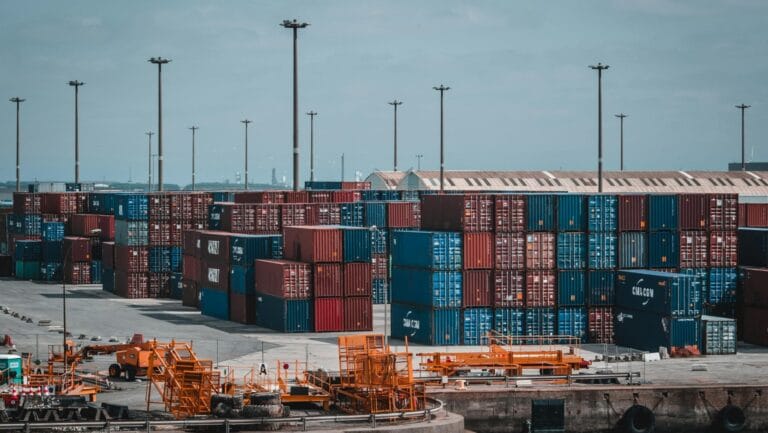 Busy shipping port with stacked red, blue, and brown cargo containers, industrial equipment, and warehouse in background